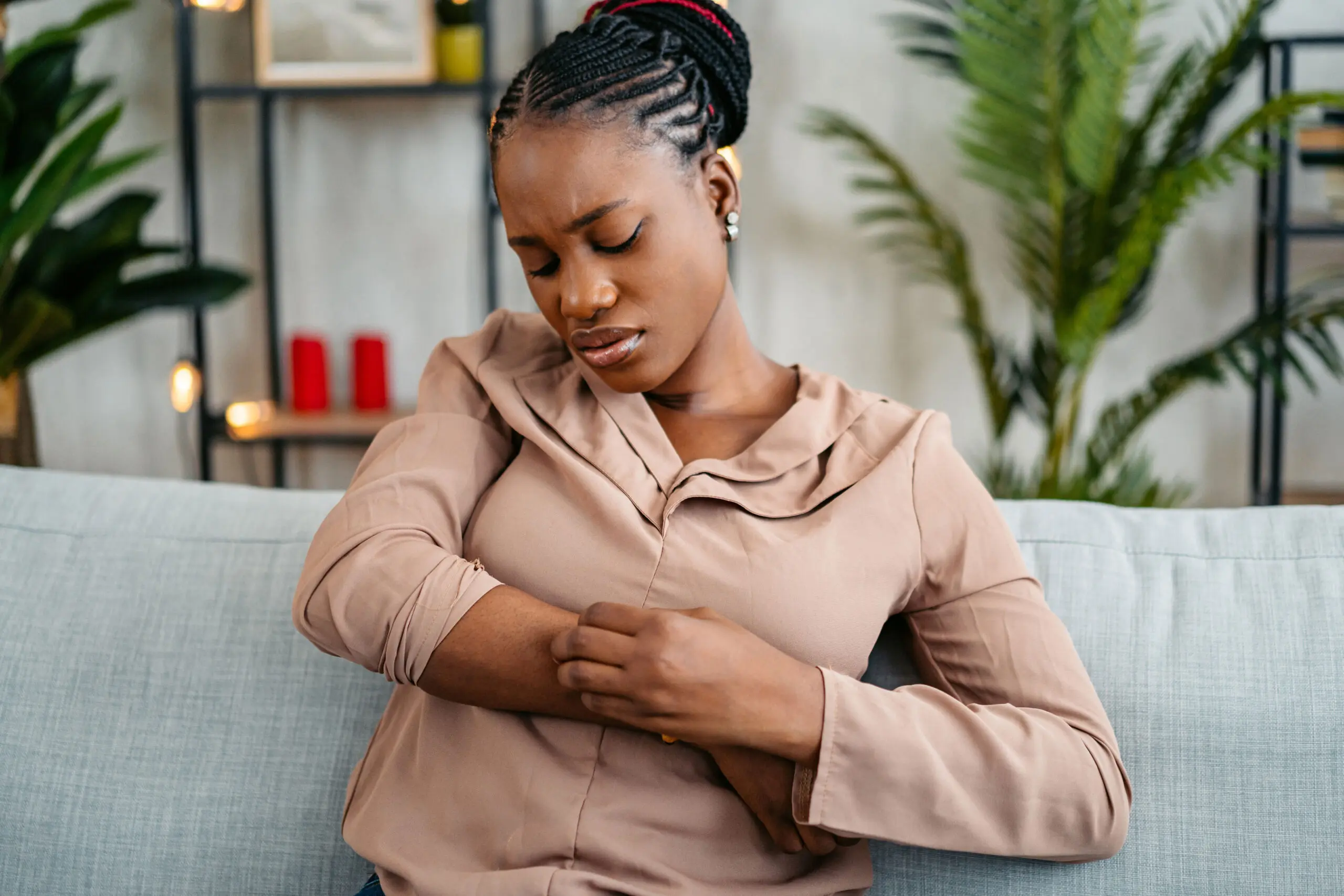 Woman with eczema scratching her arm while sitting on a couch, representing the need for fast, online eczema treatment.