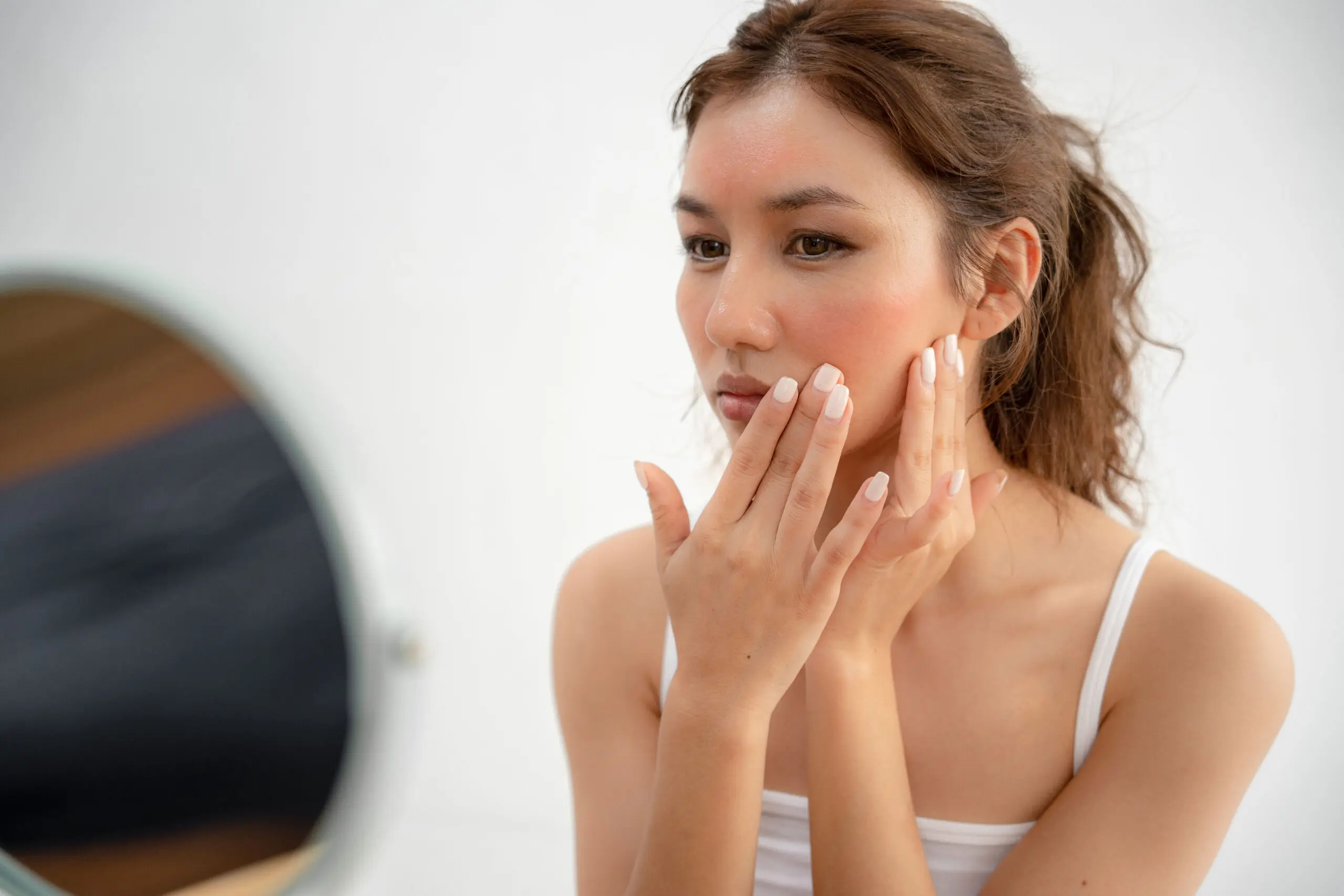 Woman examining facial skin for rash, representing when to seek dermatologist evaluation for unexplained skin symptoms.