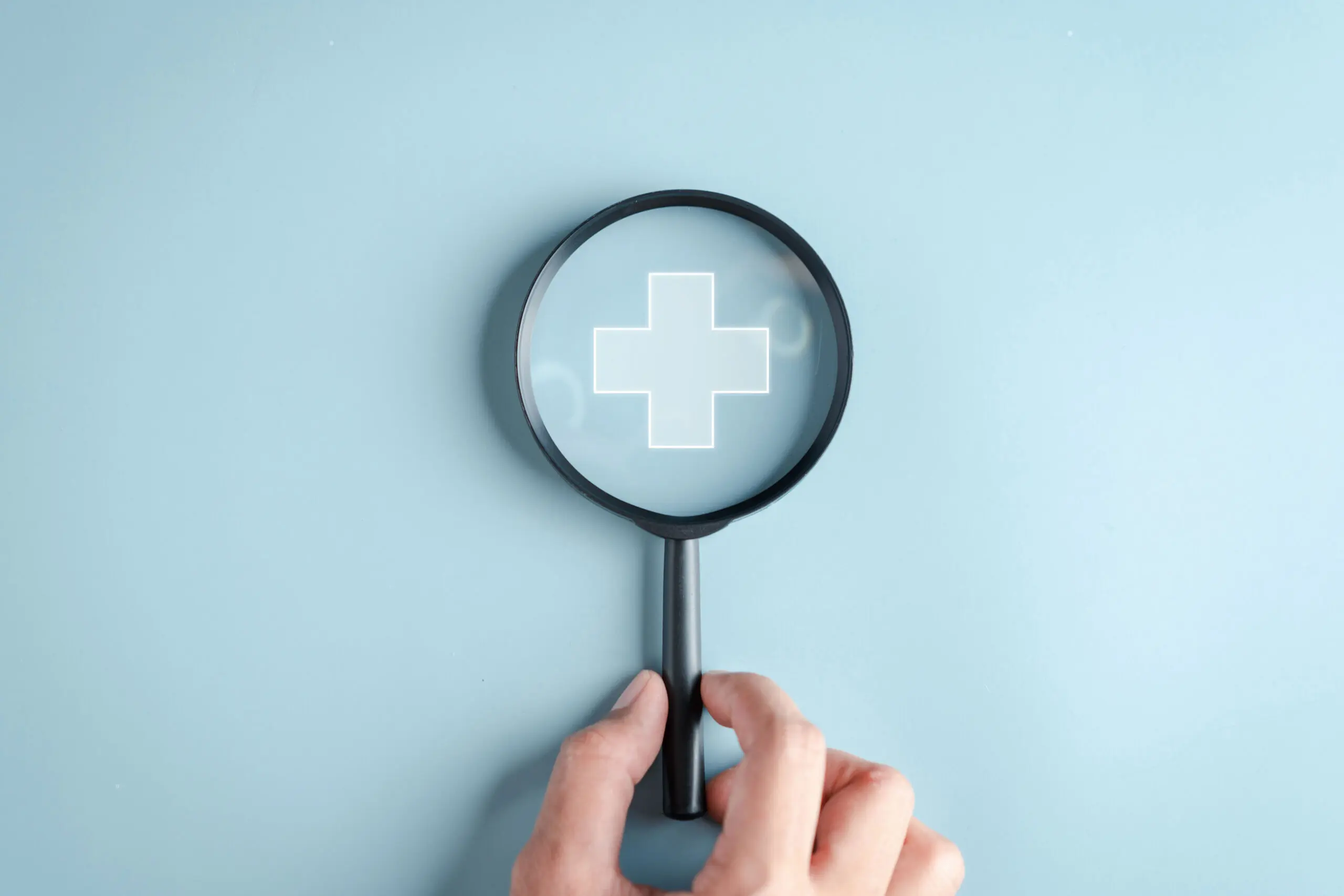 Hand holding magnifying glass over medical cross icon on blue background, symbolizing access to online dermatology without insurance.